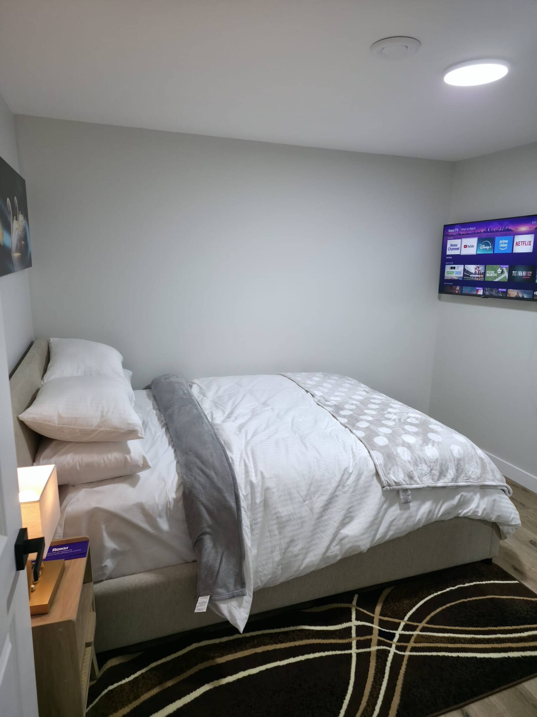 Una cama bien hecha con una colcha estampada, un televisor montado en la pared mostrando servicios de streaming y una mesita de noche con una lámpara.