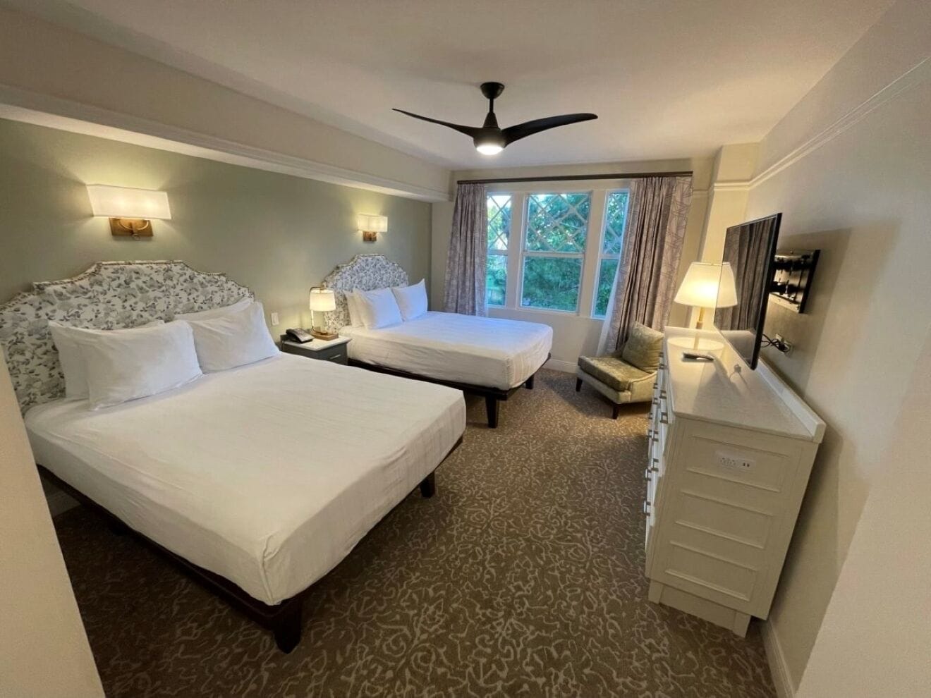 Habitación de hotel con dos camas, ventilador de techo, escritorio con lámpara y ventana con cortinas.
