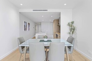 Uma sala de jantar moderna com uma mesa de vidro e quatro cadeiras, cozinha ao fundo, e um vaso de planta à direita.