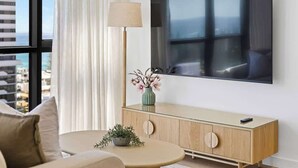 A modern living room with a large flat-screen TV mounted on the wall, a round coffee table, a sofa, a cabinet with glass doors, and a floor lamp. - 2 bed  2 bath Pool Car Park (Mermaid Beach)