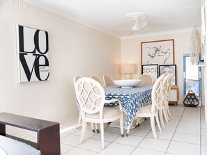 A dining area with a table set for four, white chairs, a ceiling fan, and wall art. - Mykonos Absolute Beachfront Apt Parking Ocean Views Pets Welcome (Surfers Paradise)