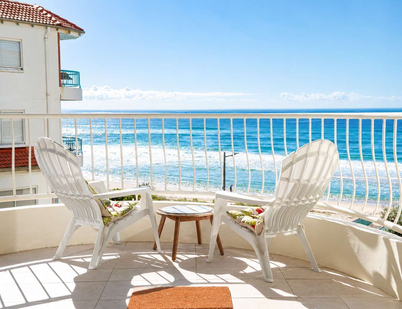 A balcony with two white chairs and a small table, overlooking a beach with the ocean and a clear sky.