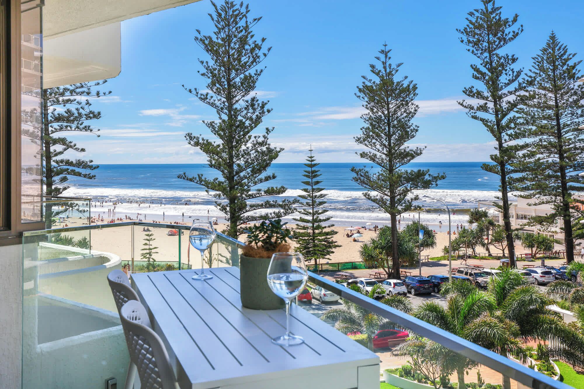 A balcony with a table and chairs, overlooking a beach with trees and a view of the ocean.