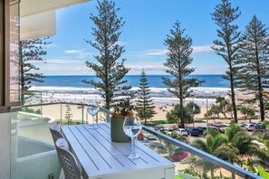 A balcony with a table and chairs, overlooking a beach with trees and a view of the ocean. - 3BR Opposite Burleigh Beach  Pool  Car Park (Burleigh Heads)