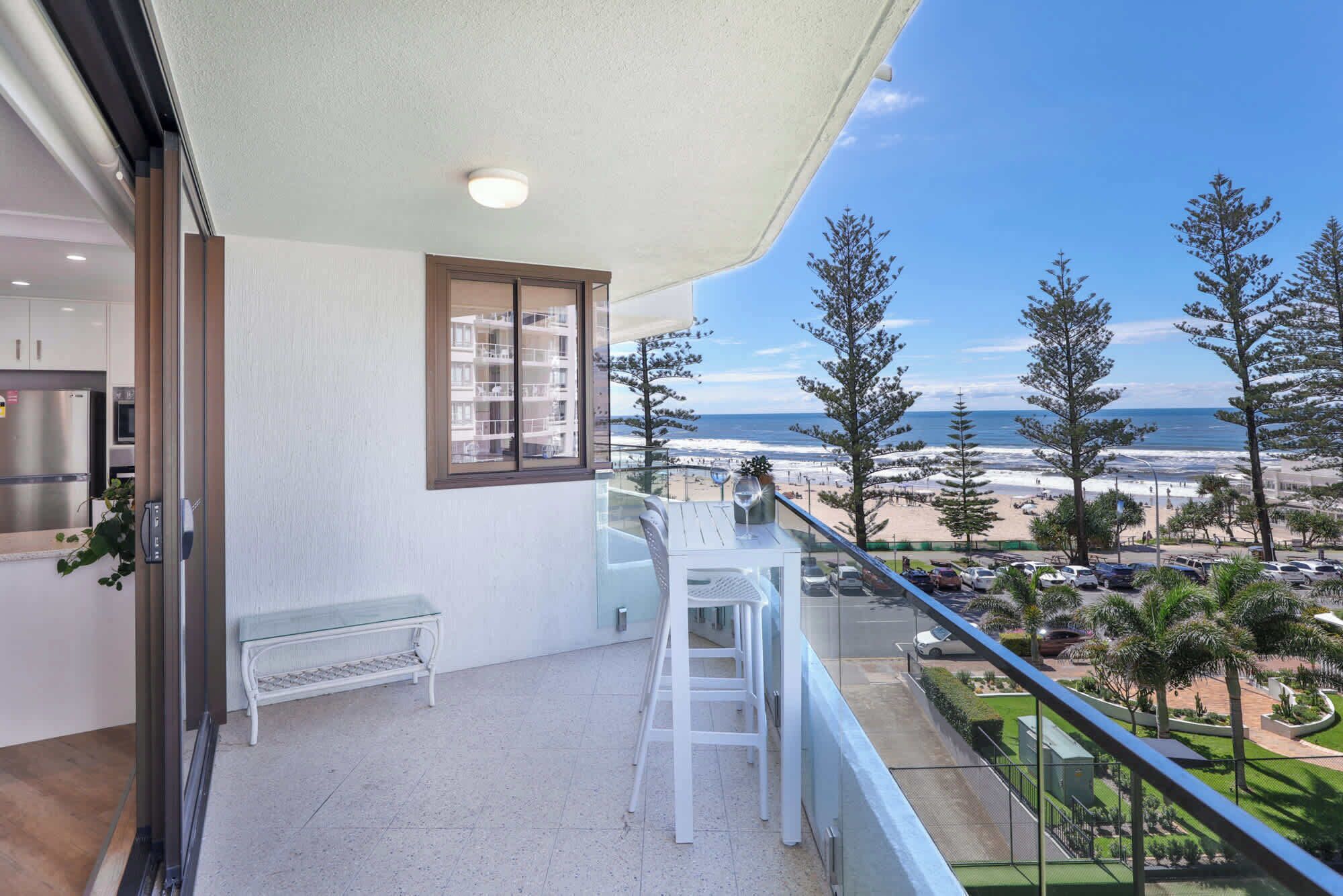 A balcony with a view of the beach, a bench, and a table with a glass.