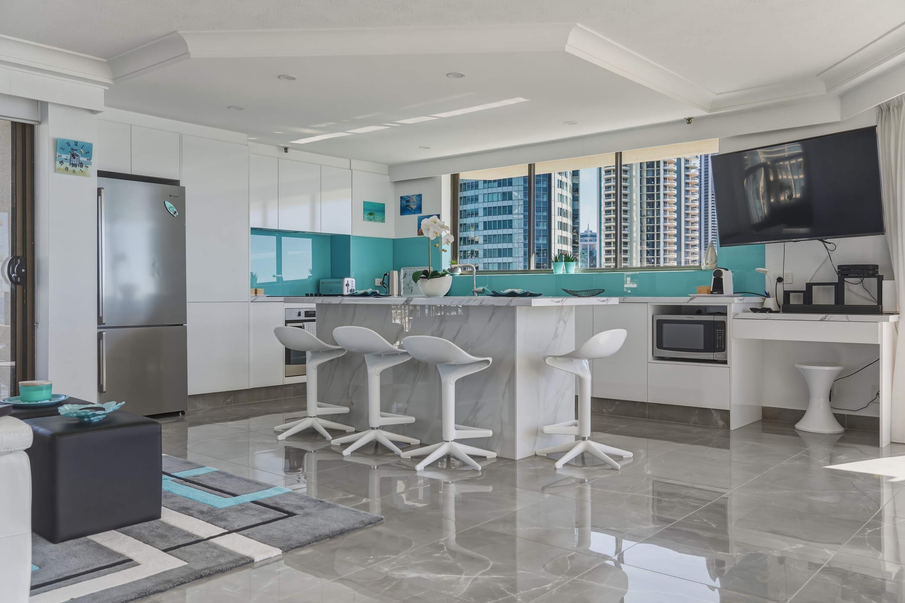 A modern kitchen with a central island, bar stools, a refrigerator, and a flat-screen TV.