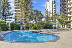 Una piscina con borde de azulejos, rodeada de sillas de descanso y una zona de estar cubierta, con edificios residenciales altos al fondo.