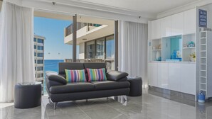A modern living room with a black leather sofa, a glass coffee table, and a view of the sea through large windows. - Lvl 15 Sunrise + Sunset Views | Pool + Parking (Surfers Paradise)