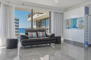 A modern living room with a black leather sofa, a glass coffee table, and a view of the sea through large windows. - Lvl 15 Sunrise + Sunset Views | Pool + Parking (Surfers Paradise)