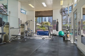 Un gimnasio con diversos equipos de ejercicio, amplias ventanas y vistas a edificios.