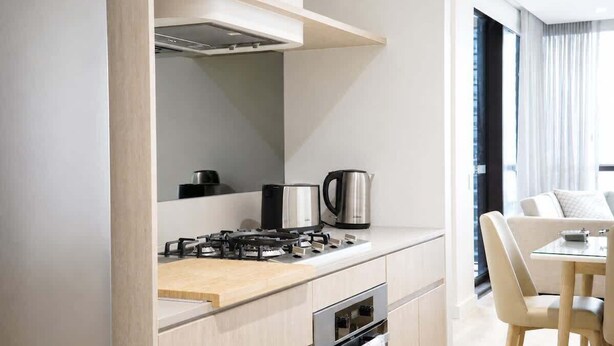 A modern kitchen with a gas stove, kettle, and oven, leading to a dining area with a table and chairs. - 2BR Ocean Views Pool Gym Sauna (Mermaid Beach)