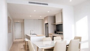A modern kitchen with white cabinetry, a central island, and a dining area with a white table and chairs. - 2BR Ocean Views Pool Gym Sauna (Mermaid Beach)