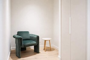Une pièce avec un fauteuil recouvert d’un tissu vert, un petit tabouret rond en bois et un mur blanc.