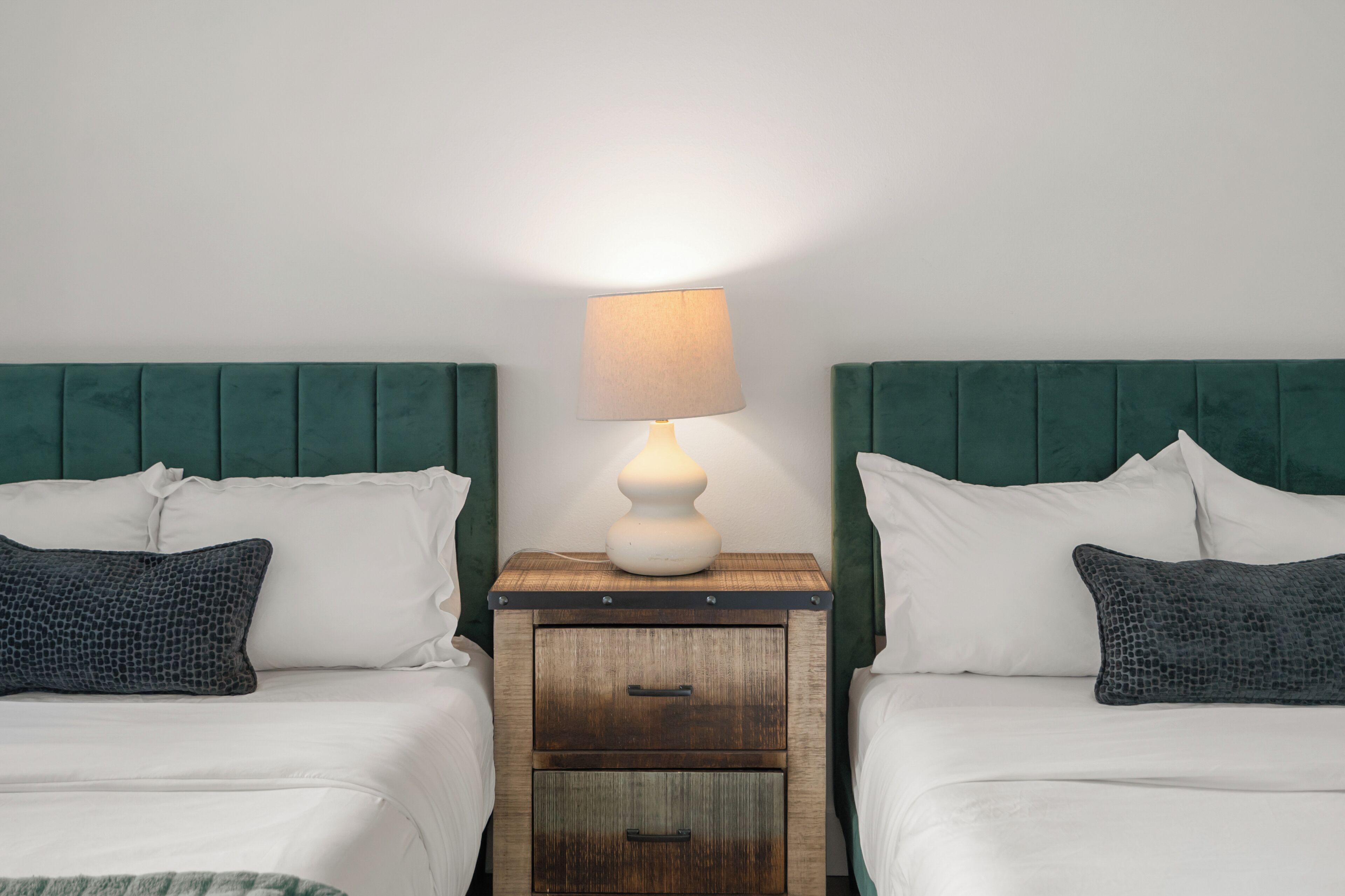 Une chambre avec deux lits, chacun doté d’une tête de lit verte, d’une table de chevet en bois avec une lampe et d’un petit tiroir.