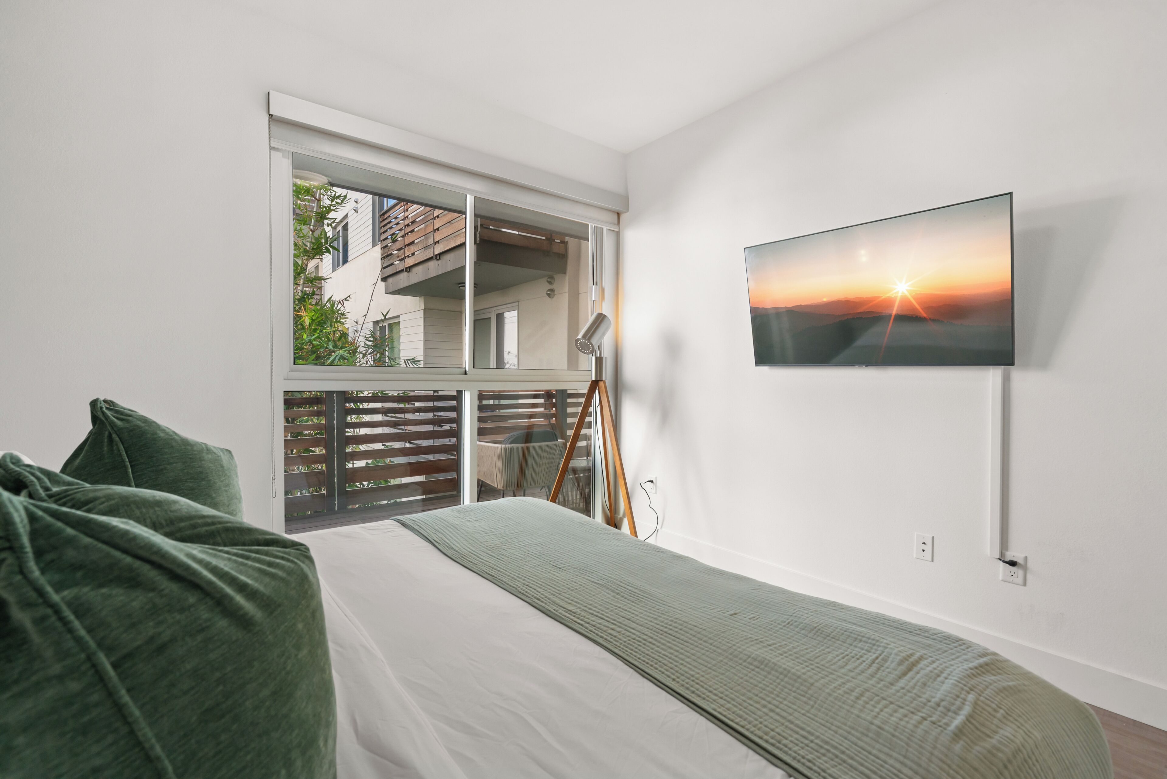 A bedroom with a bed, a TV mounted on the wall, and a window overlooking a balcony.