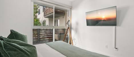 A bedroom with a bed, a TV mounted on the wall, and a window overlooking a balcony.