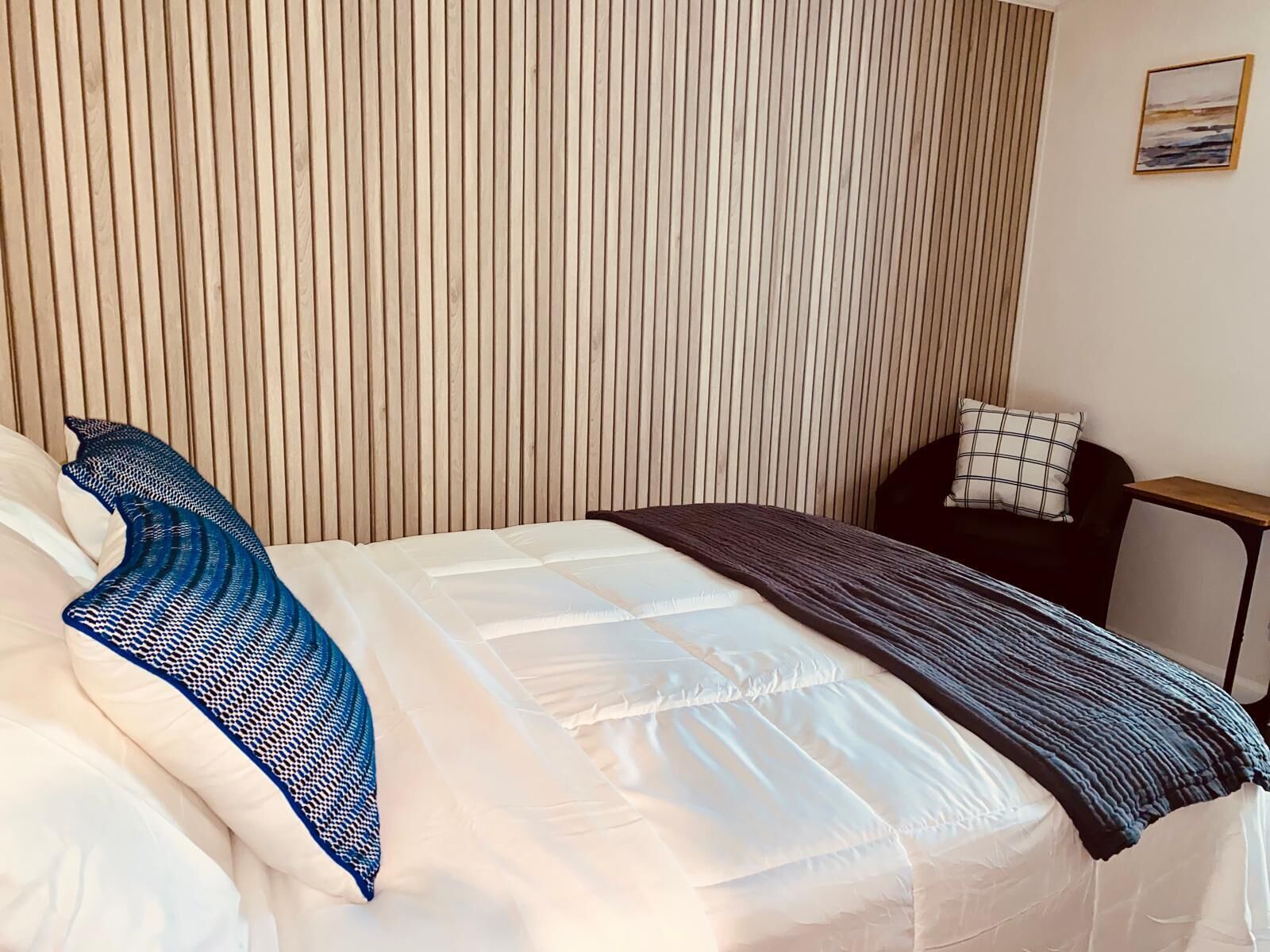 Un lit bien fait, agrémenté d’un coussin à motifs et d’une couverture, une petite table avec un coussin, et un mur avec des lattes de bois verticales.