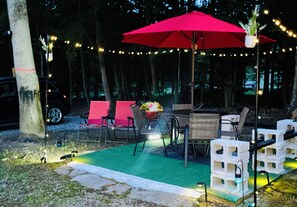 Un espace extérieur aménagé avec des sièges, un parasol rouge, des guirlandes lumineuses et une pelouse verte artificielle.