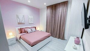 Un dormitorio con una cama, mesitas de noche, un aparador, un televisor y una ventana con cortinas.