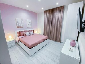 Ein Schlafzimmer mit Bett, Nachttischen, einem Schminktisch, einem Fernseher und einem Fenster mit Vorhängen.