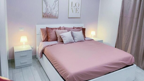 Un dormitorio con cama, dos mesitas de noche, una mesita auxiliar, una pared con dos cuadros enmarcados y una cortina.