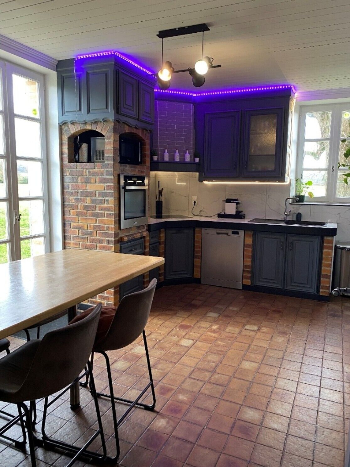 A kitchen with a brick wall, a built-in oven, a sink, and a dining area with chairs.