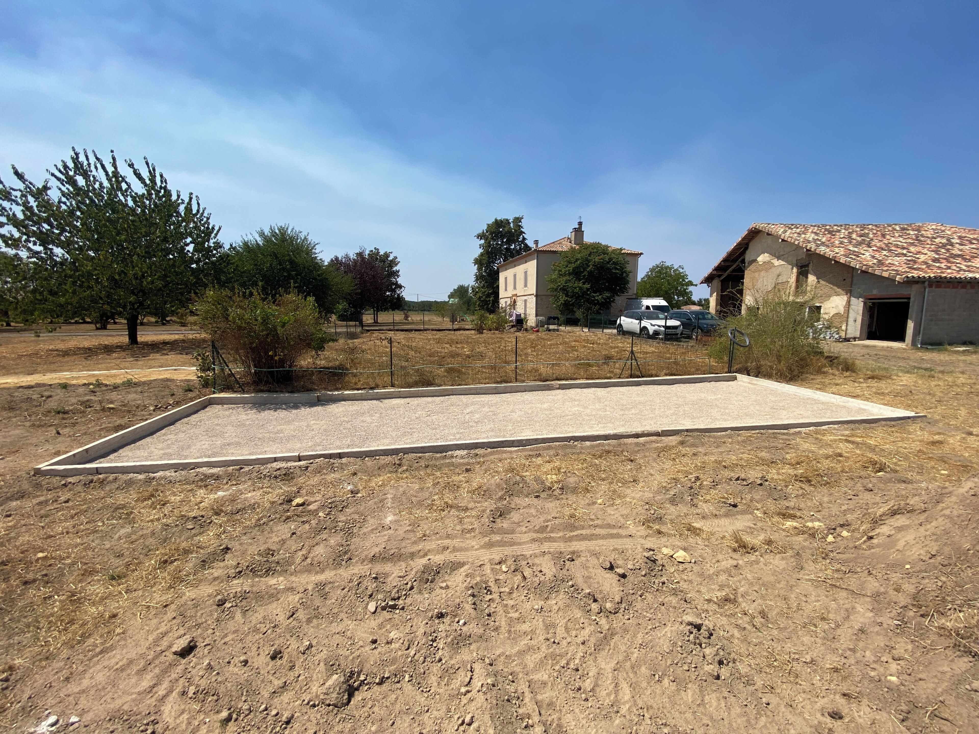 Un campo seco y despejado con una zona pavimentada, rodeado de árboles y edificios con techos de tejas.