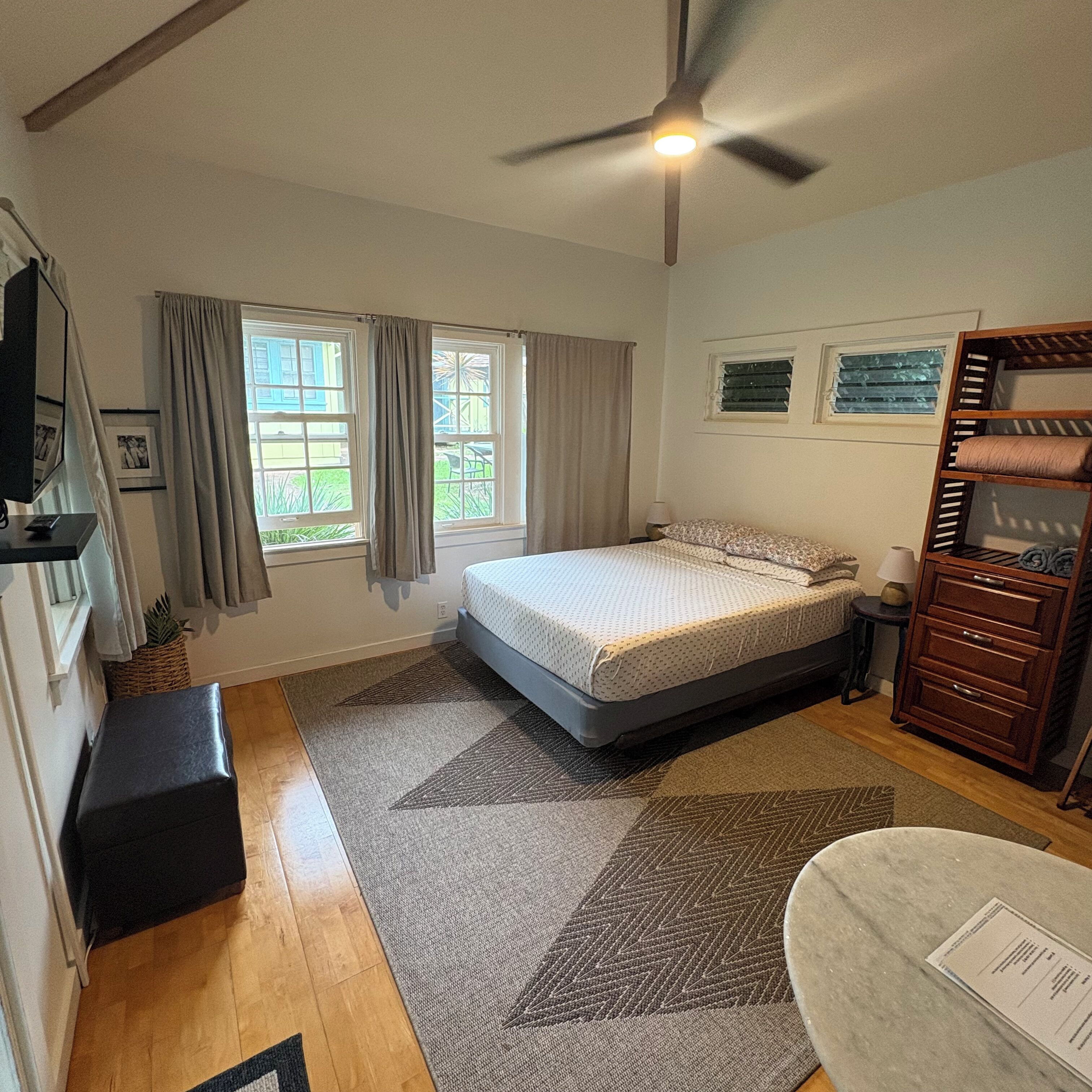 A bedroom with a bed, a ceiling fan, a window with curtains, a bedside table, a dresser, and a small rug.