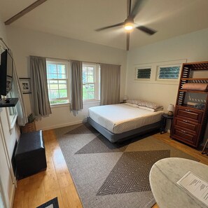 A bedroom with a bed, a ceiling fan, a window with curtains, a bedside table, a dresser, and a small rug. - Historic 5-Bedroom Home in Upcountry Maui (Makawao)