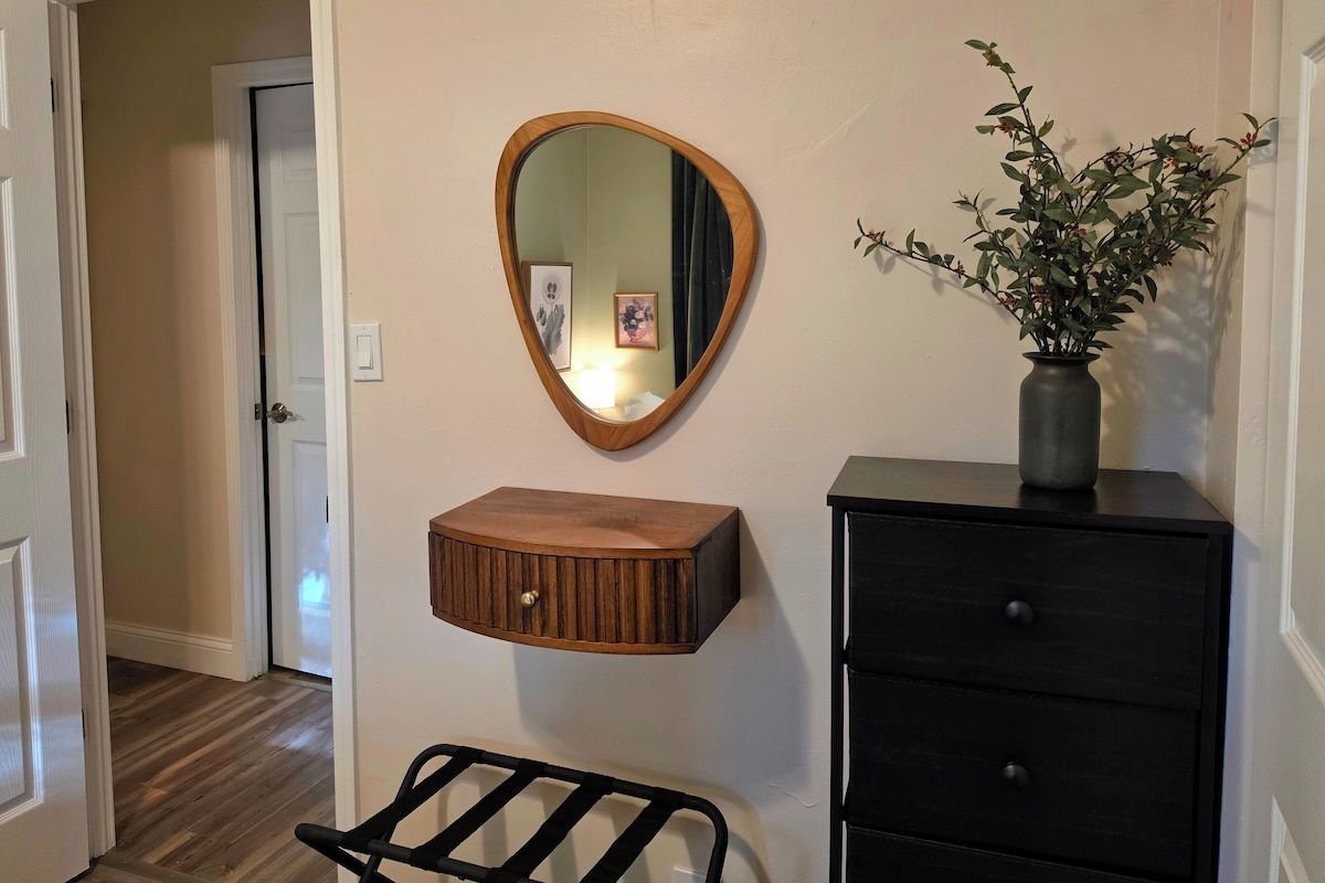 A hallway with a wooden mirror, a small wooden cabinet, a black chair, and a black dresser with a vase of flowers.
