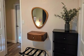 Un couloir avec un miroir en bois, une petite armoire en bois, une chaise noire et une commode noire ornée d’un vase de fleurs.