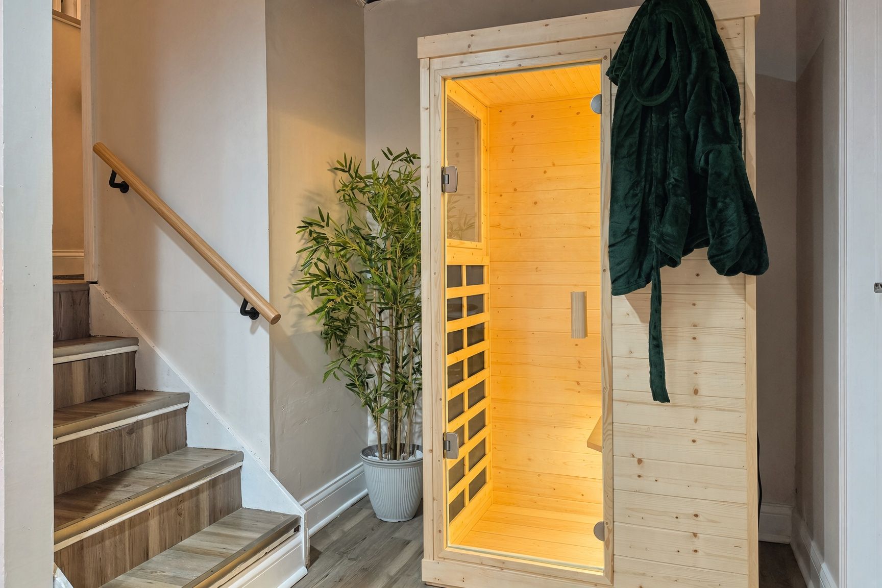 Un escalier en bois avec une rampe, un pot de bambou et une porte en bois sur laquelle est accrochée une robe verte, sur le côté droit.