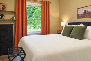 Une chambre à coucher avec un lit, une table de chevet, une lampe, une commode et une fenêtre avec des rideaux.