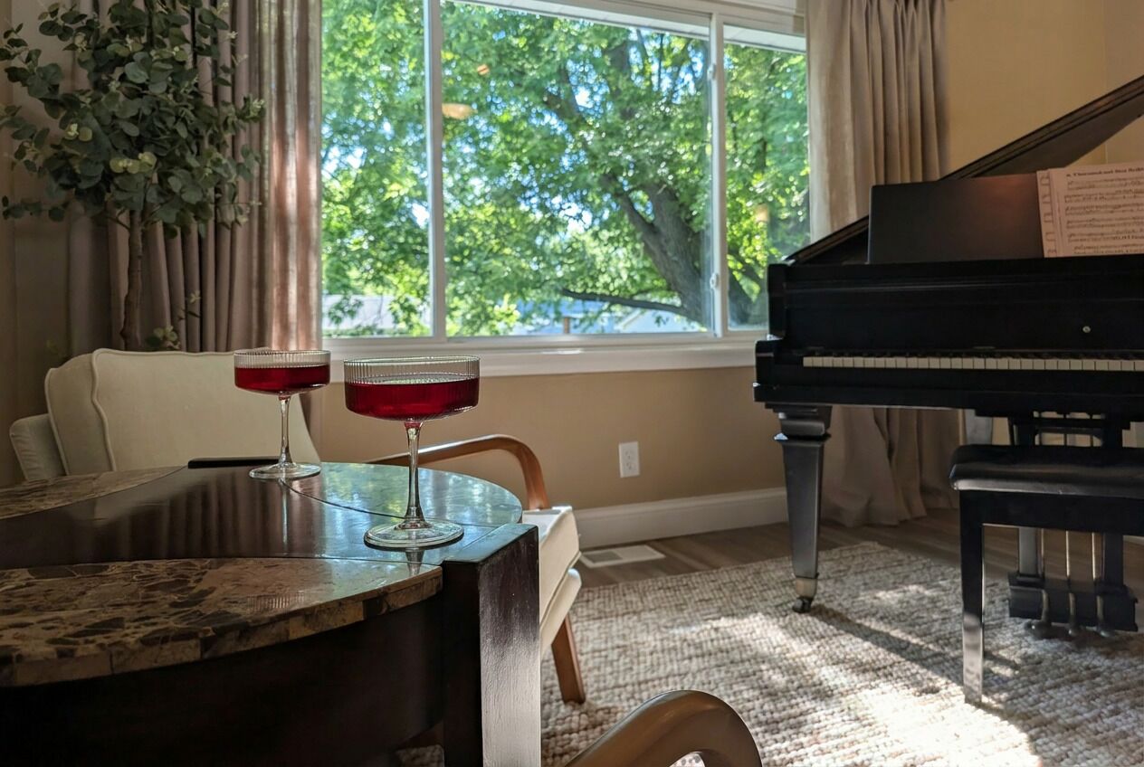 A room with a piano, a dining table with two red glasses, a plant, and a window with a view of trees.
