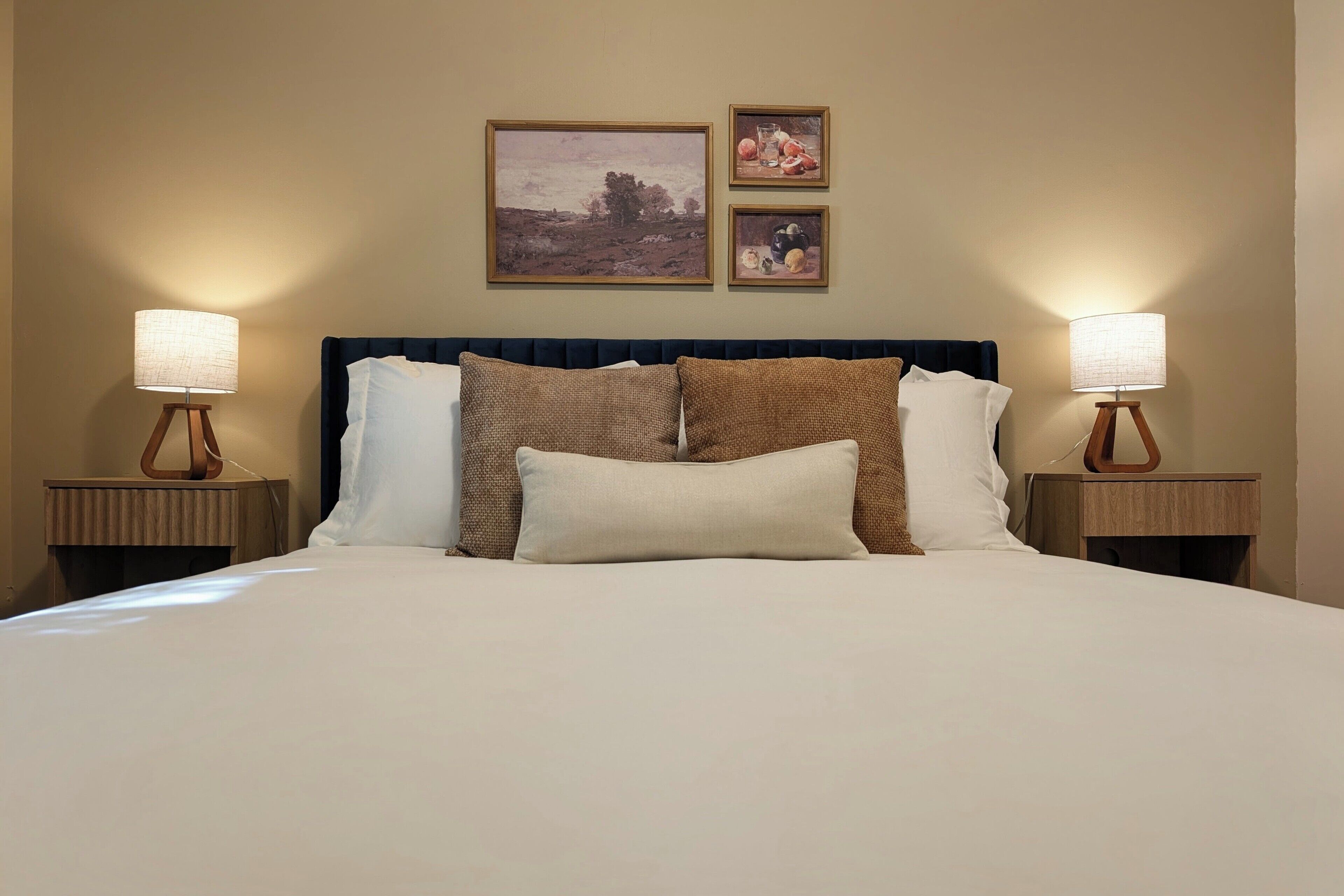 A neatly made bed with a dark headboard, two bedside tables with lamps, and two framed artworks above the bed.