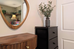 A wooden vanity with a mirror, a vase with green foliage, and a black cabinet with drawers.