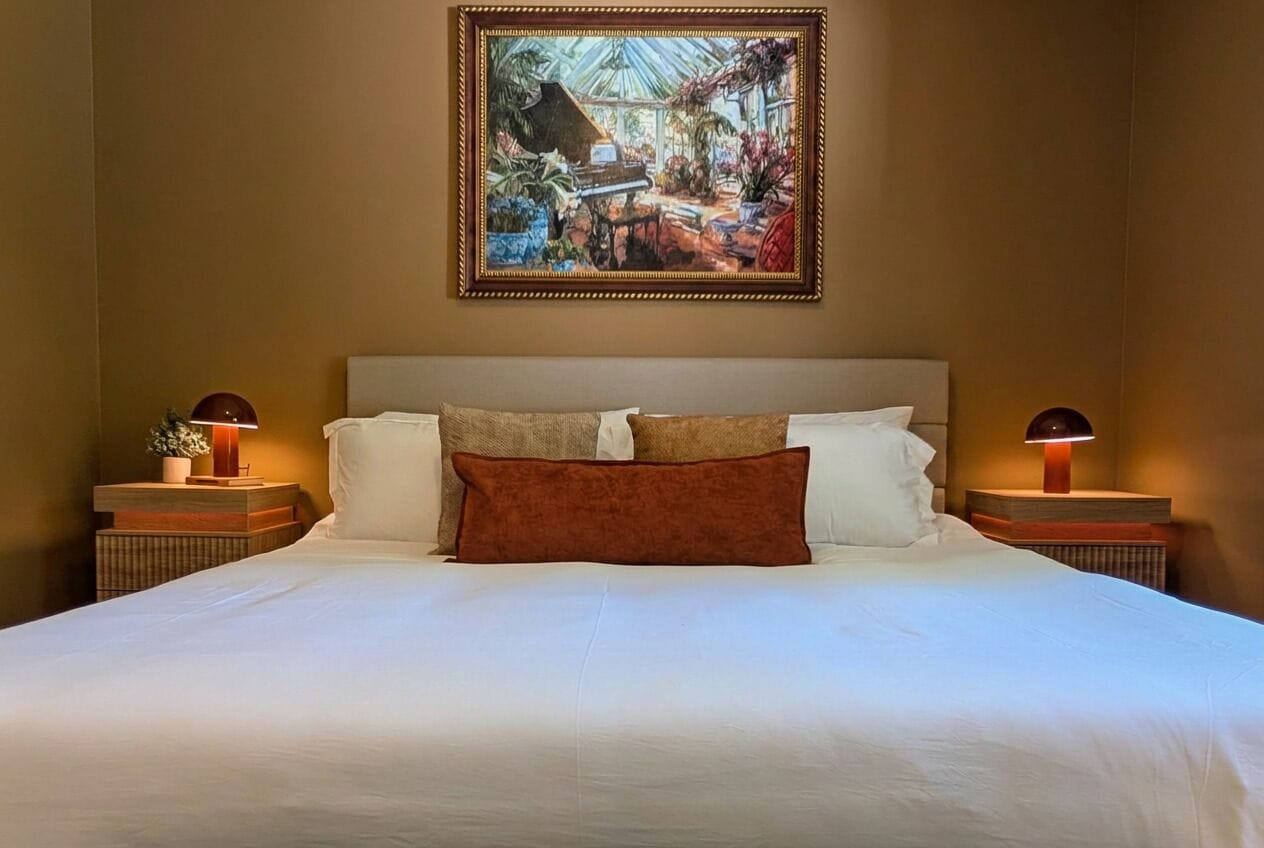 Quarto de hotel com uma cama grande, duas mesas de cabeceira, um quadro emoldurado acima da cama e uma mesinha de cabeceira com abajur.