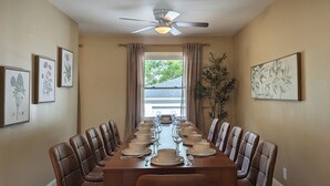 A dining room with a long table set for a meal, flanked by chairs, with ceiling fans and framed botanical prints on the walls. - La Petite • Piano • Serene Organic • Long Stays (Merrillville)