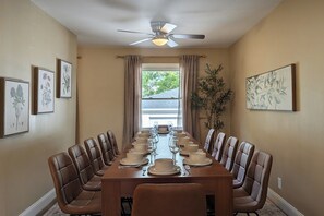 A dining room with a long table set for a meal, flanked by chairs, with ceiling fans and framed botanical prints on the walls.