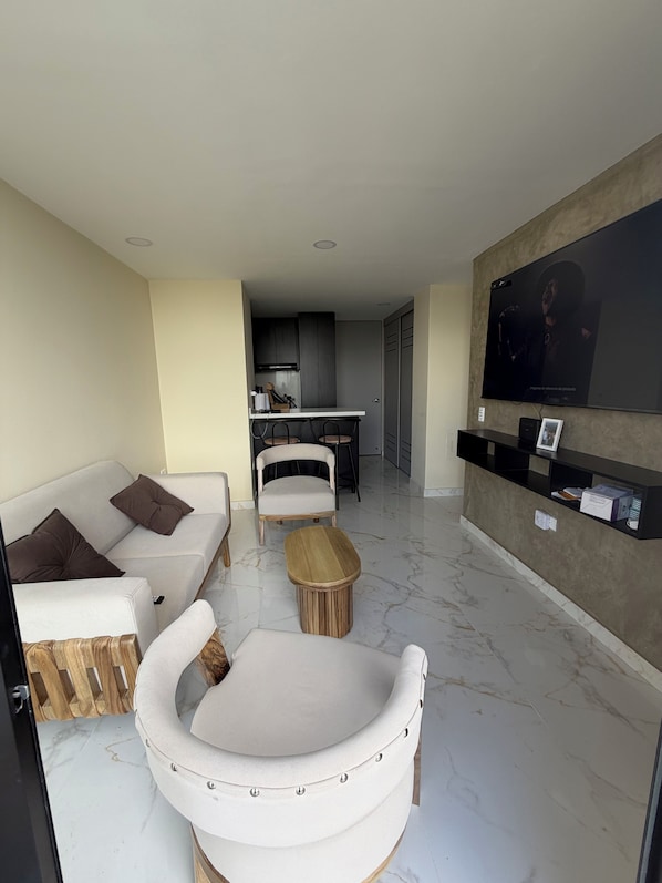 A modern living room with a sofa, armchair, and coffee table. A kitchen is visible in the background.