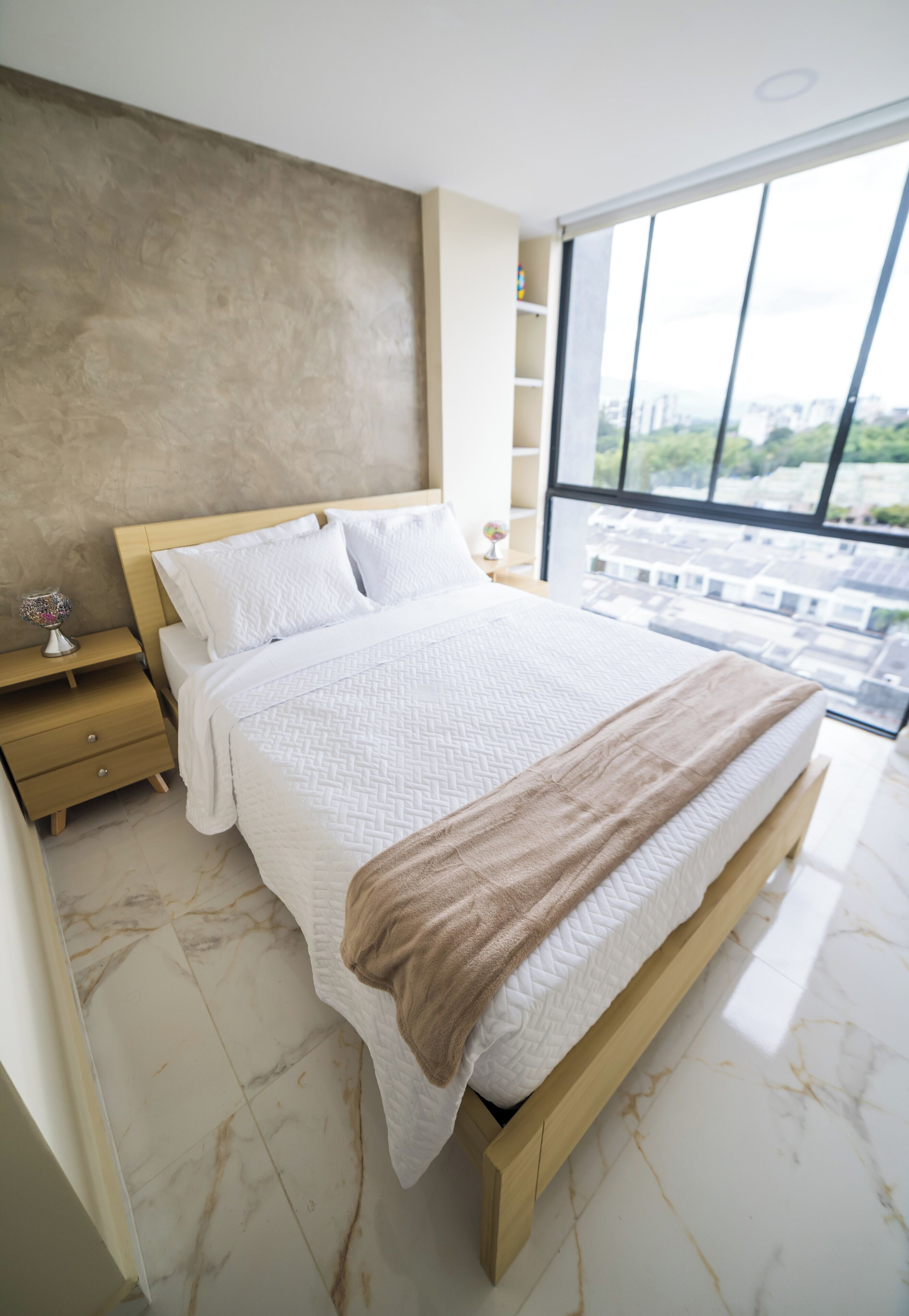 A modern bedroom with a large bed, a nightstand, a globe, and a view of the city through large windows.
