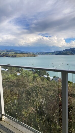 A coastal view from a balcony with a railing, overlooking a calm sea with boats, and a hilly landscape in the distance.