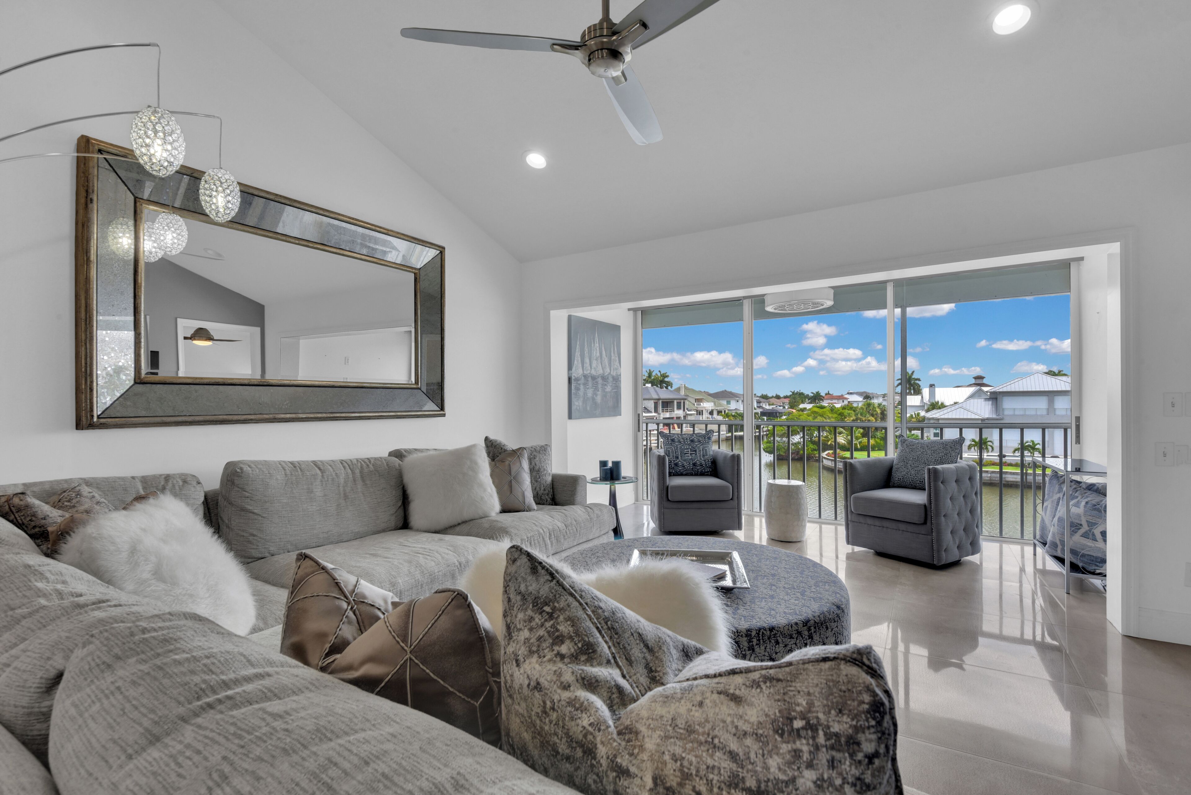 A modern living room with a large sofa, a ceiling fan, and a view of a body of water through large glass doors.