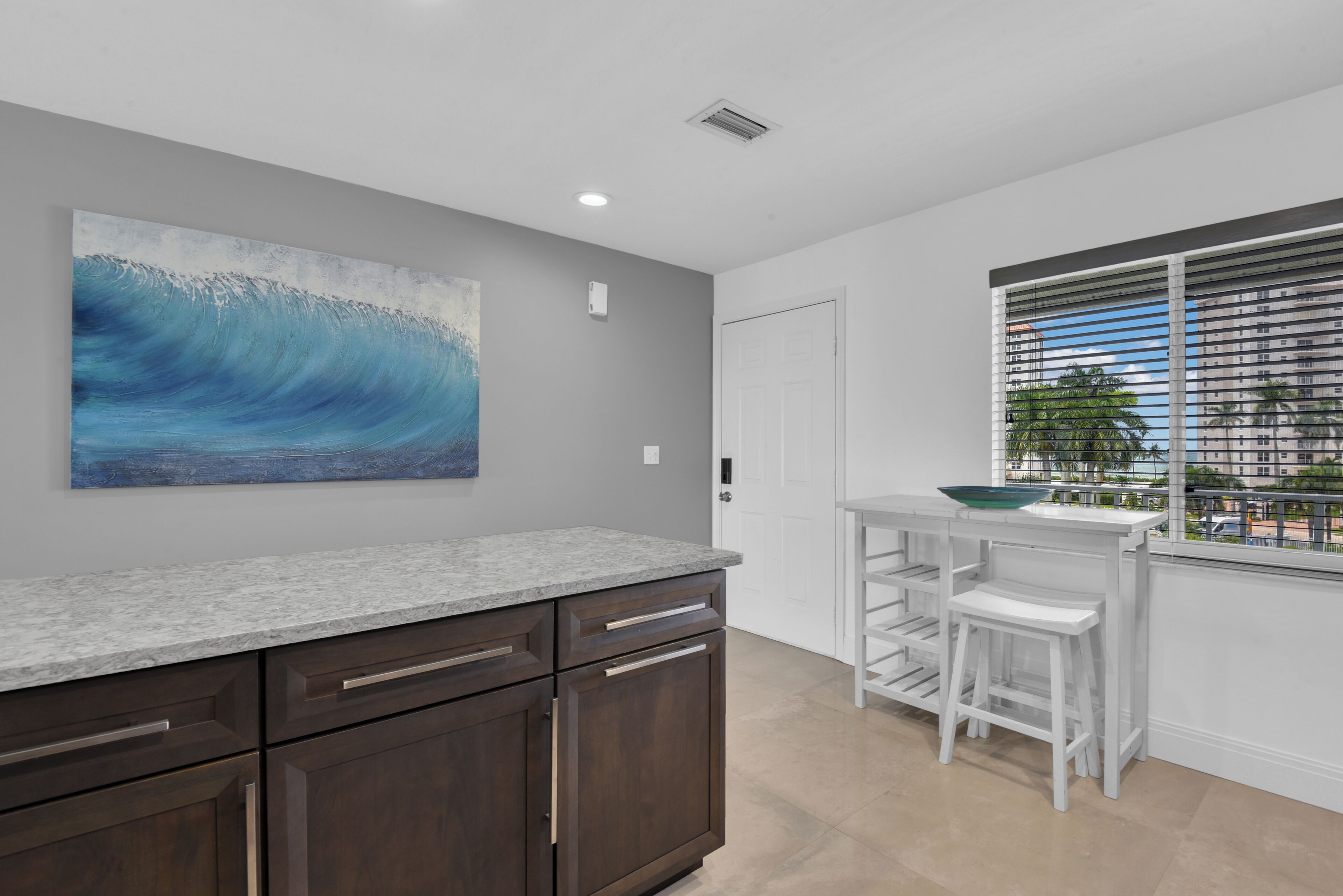 A kitchen with a granite countertop, dark wooden cabinets, a white bar stool, and a window with blinds.
