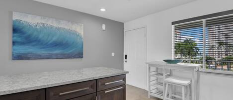 A kitchen with a granite countertop, dark wooden cabinets, a white bar stool, and a window with blinds.