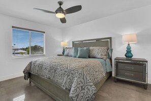 Une chambre à coucher comprenant un lit, une table de chevet, une lampe, un ventilateur de plafond et une fenêtre avec des stores.