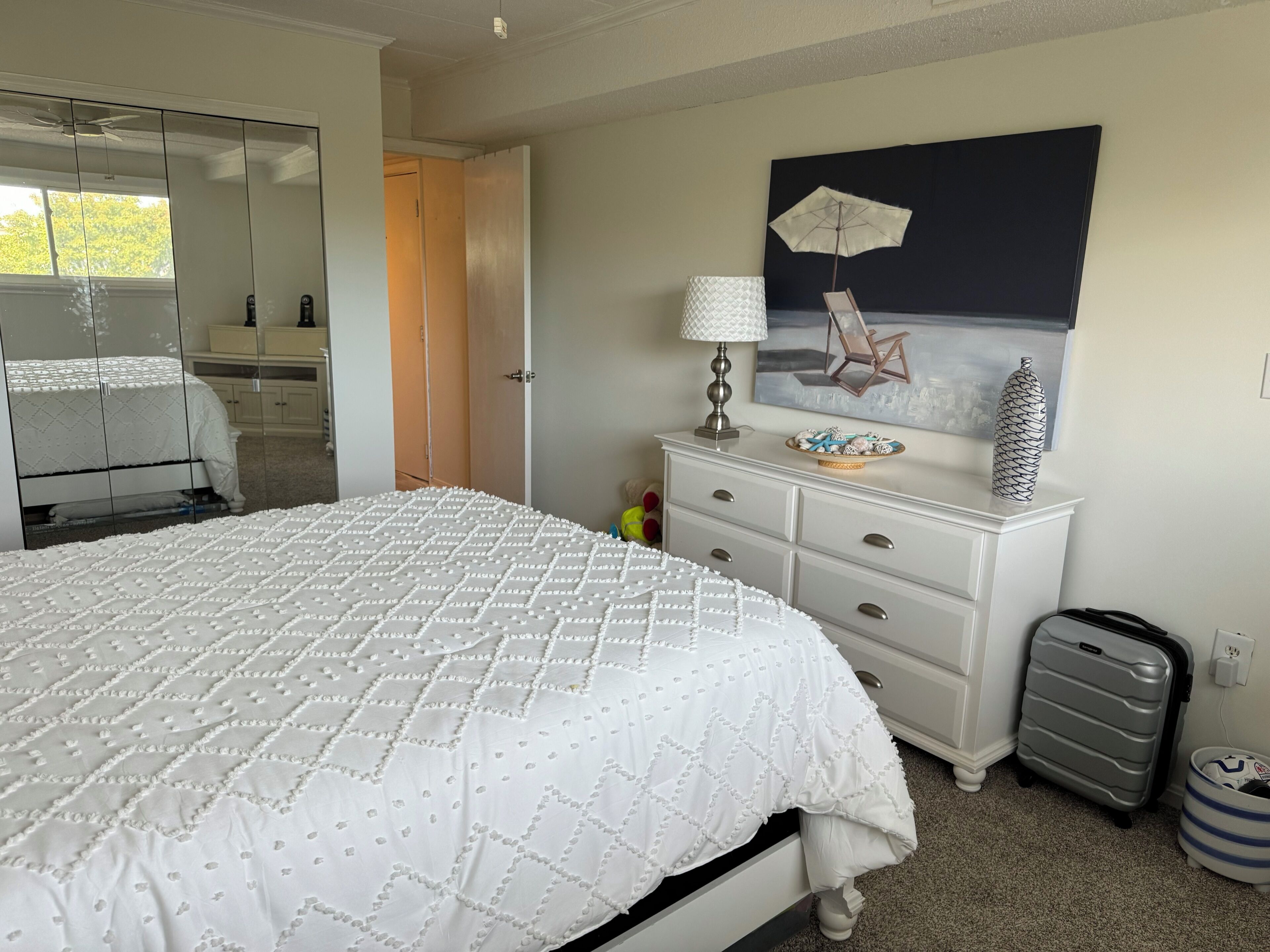 Ein Schlafzimmer mit Bett, Kommode, Lampe, Koffer und einem Bild an der Wand.