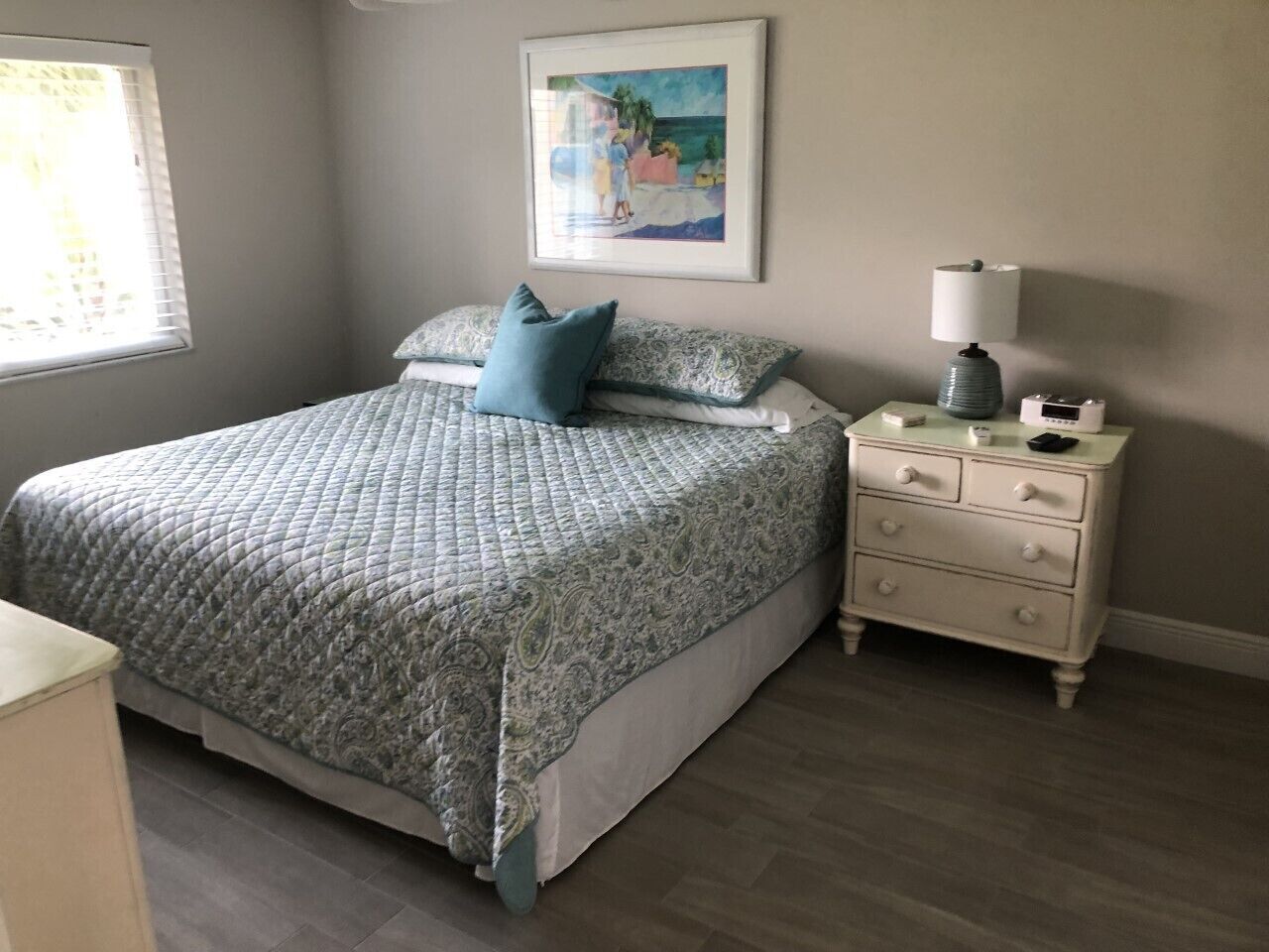 Un dormitorio con cama, mesita de noche, lámpara y un cuadro en la pared.