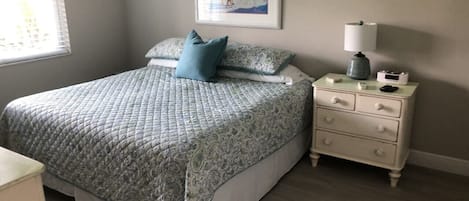Un dormitorio con cama, mesita de noche, lámpara y un cuadro en la pared.
