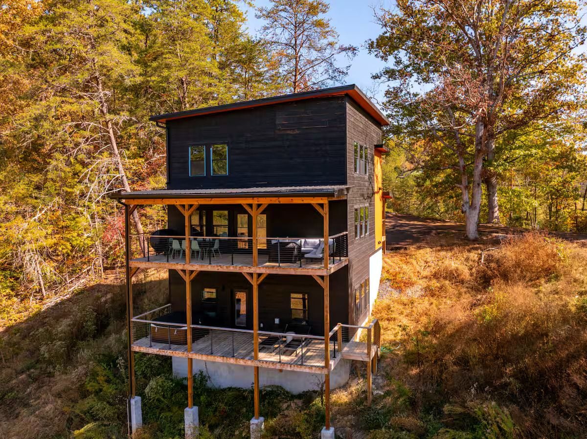 A multi-story cabin with a balcony on the ground floor, surrounded by trees.